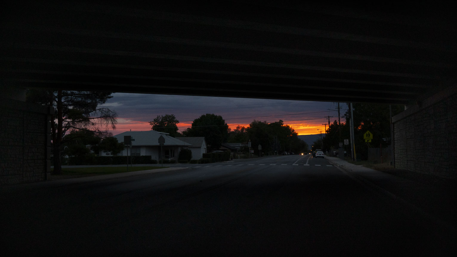 The sunset blazes in the distance behind trees and houses all seen through the rectangle opening of the underpass